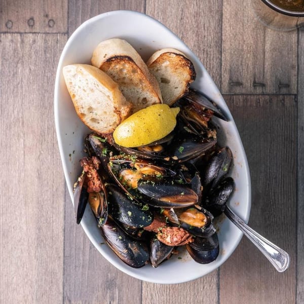 A white bowl of mussels with chorizo, toasted bread, and a lemon wedge on a wooden table.