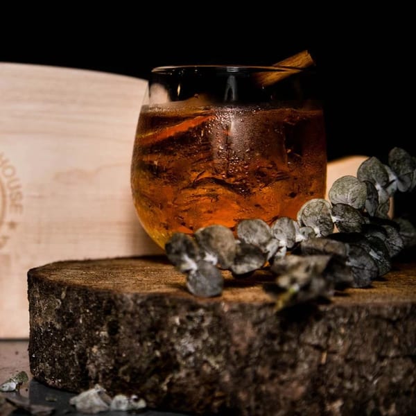 A Wildfire Public House cocktail with ice and a cinnamon stick, garnished with dried eucalyptus on a wood slab.