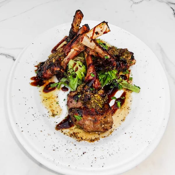 Grilled lamb chops with a dark glaze, green herbs, and a side of fresh greens on a white plate.