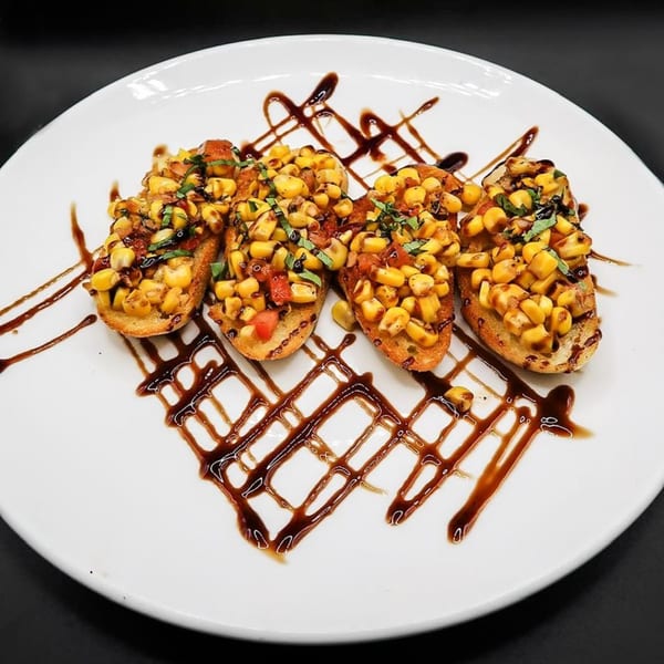Four corn and tomato bruschetta pieces drizzled with balsamic glaze on a white plate.