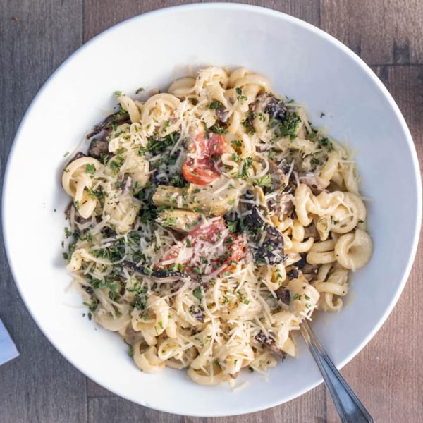 Creamy pasta with mushrooms, artichoke hearts, tomatoes, and parsley in a white bowl.