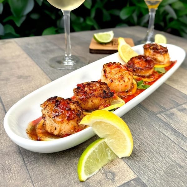 Seared scallops with red sauce, lemon, and lime wedges on a white platter, with wine glasses.